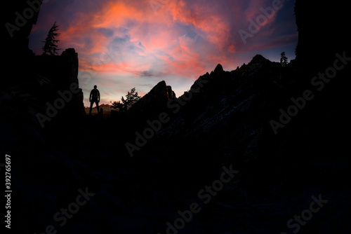 A silhouette of an adventurous male hiker standing on top of a cliff enjoying the beautiful view during a vibrant sunset. 