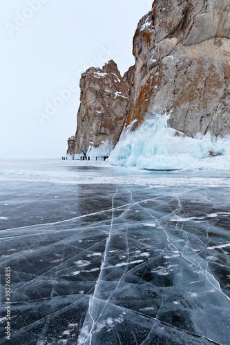 Beautiful winter landscape of frozen Lake Baikal on cold February day. Blue transparent ice with cracks near the Rocks of Olkhon Island. A group of tourists take pictures of icy rocks. Winter travel