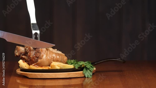 Cut off a large piece of meat from the pork knuckle with a knife. Very fatty junk food. Knuckle on a wooden background. Slow motion