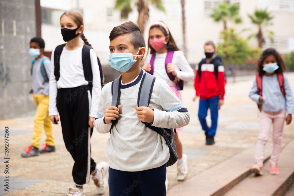 Focused hispanic tweenager in medical face mask with backpack going to school lessons on warm autumn day. New lifestyle during coronavirus pandemic.