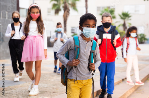 Portrait of schoolboy in medical mask standing near school, kids on background