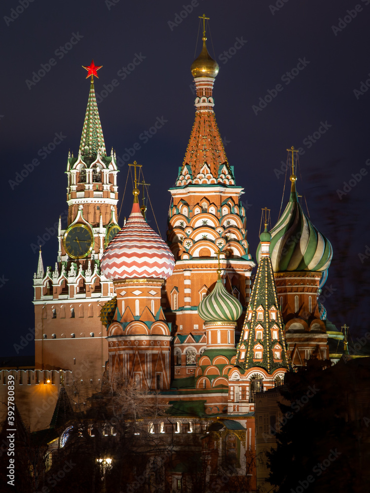Night cityscape of brightly lighted St. Basil Cathedral and Kremlin tower in Moscow. Dark blue sky. Blurred silhouette of a tree. No people.
