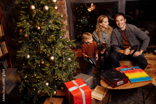 Happy parents with little son sit near Christmas tree, happy family listening music plates, smiling, enjoy festive mood, vinyl music, winter holidays at home, xmas concept