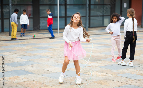 Cute tween girl in pink skirt jumping rope in school yard during recess in warm fall day.