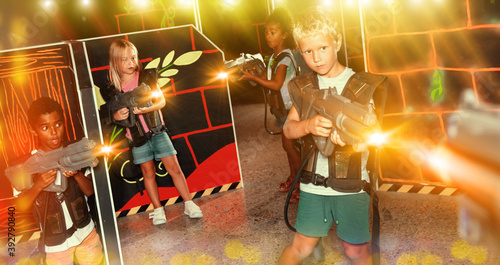 Multiracial team of happy positive smiling tween kids aiming laser guns at other players during lasertag game in dark room