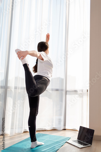 A young woman is doing yoga on a Mat. The laptop is on the floor. Windows in the background. Rear view. Training at home with online lessons