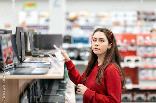 Beautiful young woman in a red sweater, choosing a laptop. In the background, shelves with electronics. Concept of choosing and purchasing digital equipment