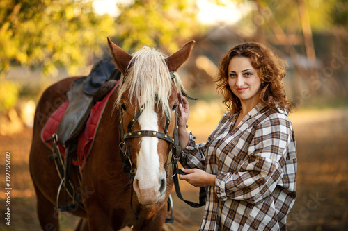 a young beautiful brunette woman in a plaid shirt holds a brown horse by the bridle