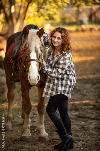 a young beautiful brunette woman in a plaid shirt holds a brown horse by the bridle