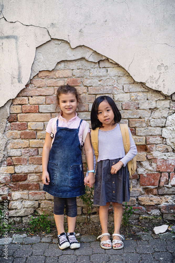 Small girls looking at camera outdoors in town, standing against old brick wall.