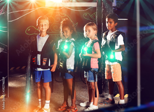 Portrait of multiracial team of smiling preteen kids with laser guns during lasertag game on dark arena