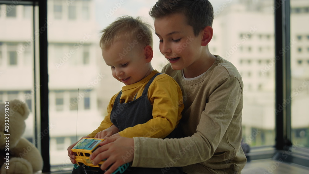 Obraz premium Younger brother sitting in older laps on carpet. Children playing toy car.
