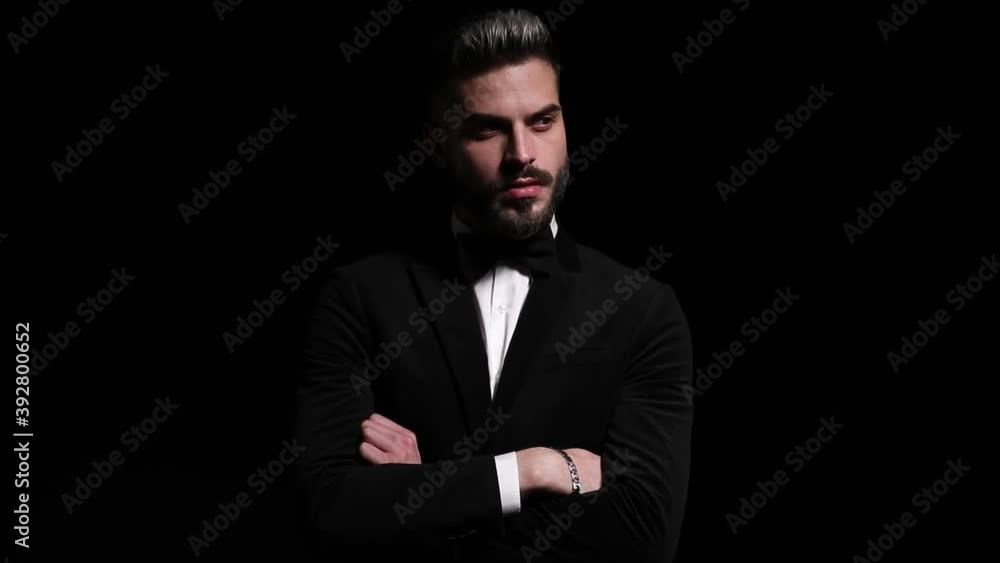 serious elegant man in black tuxedo crossing arms and looking to side, holding fingers to mouth and making silence gesture on black background in studio serious elegant man in black tuxedo crossing arms and looking to side, holding fingers to mouth and making silence gesture on black background in studio