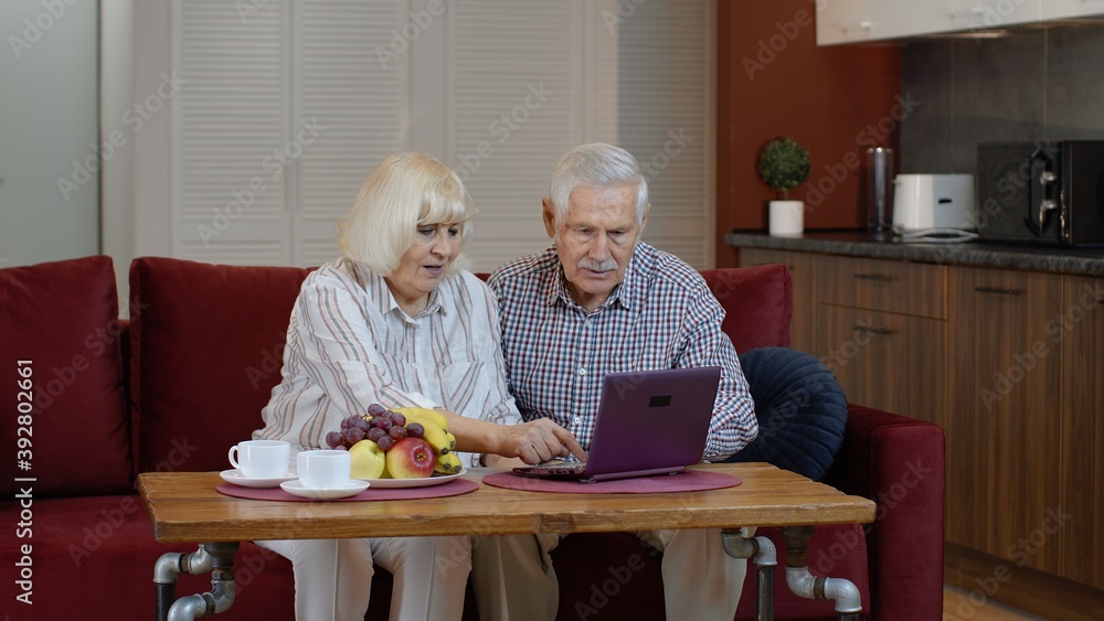 Happy smiling senior couple with laptop pc computer at home. Cheerful retired husband and wife hugging and laughing using digital computer resting on sofa in living room Happy smiling senior couple with laptop pc computer at home. Cheerful retired husband and wife hugging and laughing using digital computer resting on sofa in living room