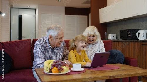 Child girl shows something in laptop to grandparents, seniors couple learning how to use computer. Grandfather and grandmother with granddaughter in living room during coronavirus quarantine lockdown