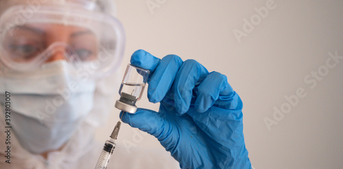 Young female doctor wearing full PPE protective gear against coronavirus covid-19 virus, pulls the plunger back to fill a syringe with a vaccine from a vial. White background. Panoramic format 