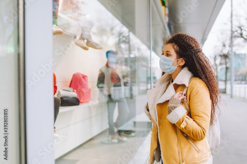 Beautiful afro haired woman wearing protective medical face mask stand on the street of city and look at shop window.