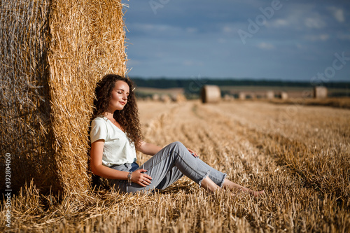 Beautiful young woman near a sheaf of hay in a field. Holidays in the village, a girl enjoying nature in a mown field on a sunny day