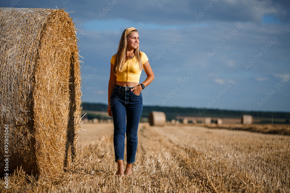 A young beautiful blonde stands on a mown wheat field near a huge sheaf of hay, enjoying nature. Nature in the village A young beautiful blonde stands on a mown wheat field near a huge sheaf of hay, enjoying nature. Nature in the village