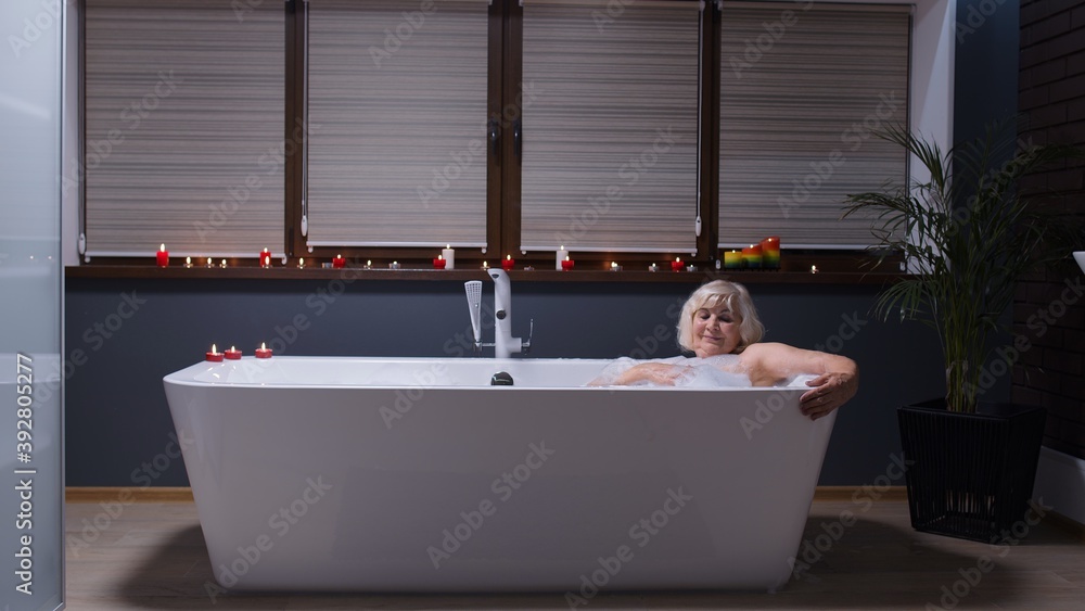 Sexy senior blonde woman grandmother is taking foamy bath in luxury bathroom with candles