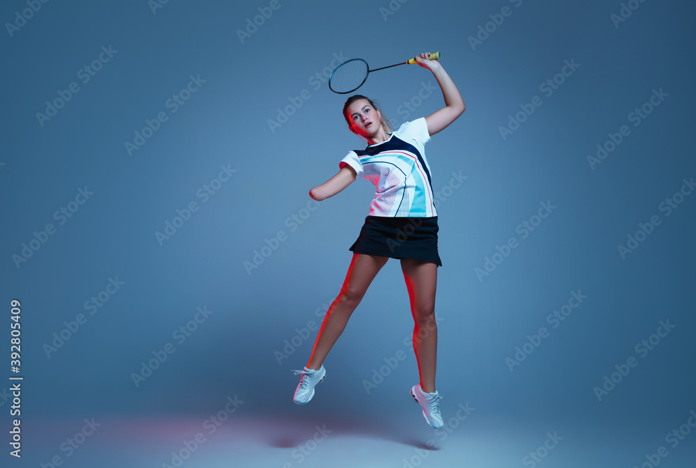In jump. Beautiful handicap woman practicing in badminton isolated on blue background in neon light. Lifestyle of inclusive people, diversity and equility. Sport, activity and movement. Copyspace.