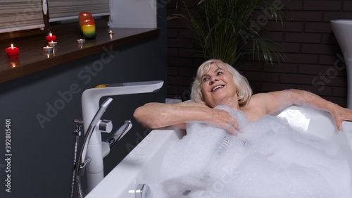 Sexy senior blonde woman grandmother is taking foamy bath in luxury bathroom with candles. Beautiful elderly lady grandma strokes skin. Spa procedures self-care. Skin care. Life of active retirees