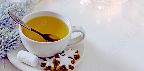 Homemade christmas tea party background. White mug of tea with asterisk cookies, dried oranges, cinnamon sticks on a white table, surrounded by new decor and garlands. Cozy winter, hygge atmosphere