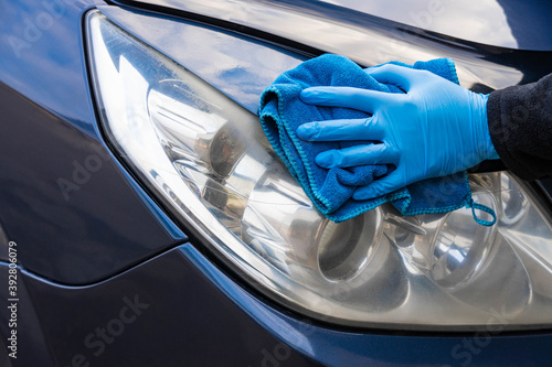 A hand in a blue rubber glove with a blue microfiber rag rubs and polishes the headlight of a car. Wash the car yourself