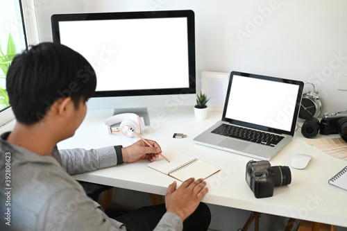 A graphic designer or photographer is taking note while sitting in front of multiple computer at workplace.