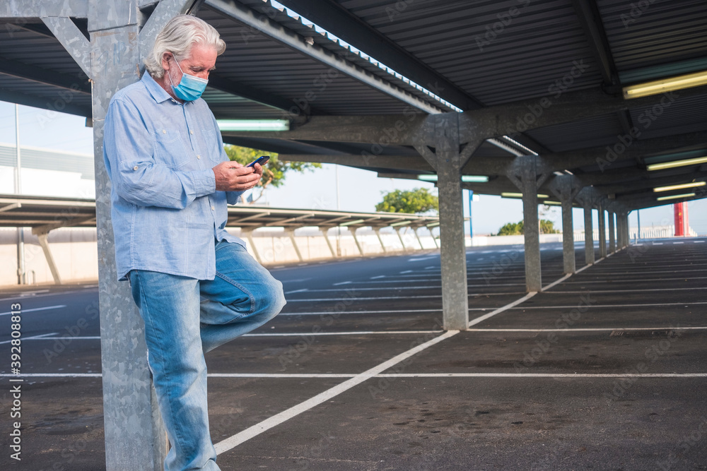 Coronavirus. A senior man wearing medical mask due to coronavirus standing under the metal structure of a deserted parking for lockdown looking at his smart phone. Nobody else