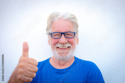 Portrait close-up of a smiling senior man with white beard on white background wearing eyeglasses and tshirt in blue color. Positive retiree person with thumb up