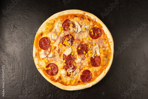 Round hot freshly baked pizza with salami, chicken, corn, onion and cheese lies on a black stone kitchen table