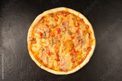 Round hot freshly baked pizza with bacon paprika pineapple and cheese lies on a black stone kitchen table