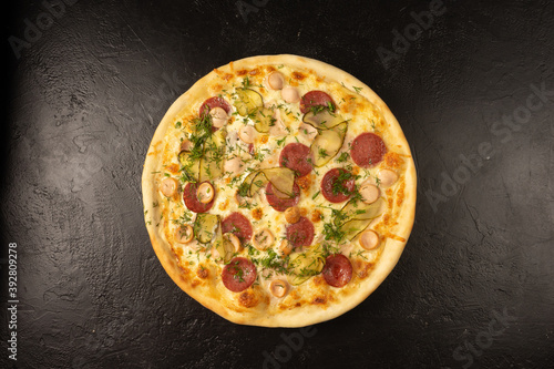 Round hot freshly baked pizza with salami, sausages, pickled cucumber, dill and cheese lies on a black stone kitchen table