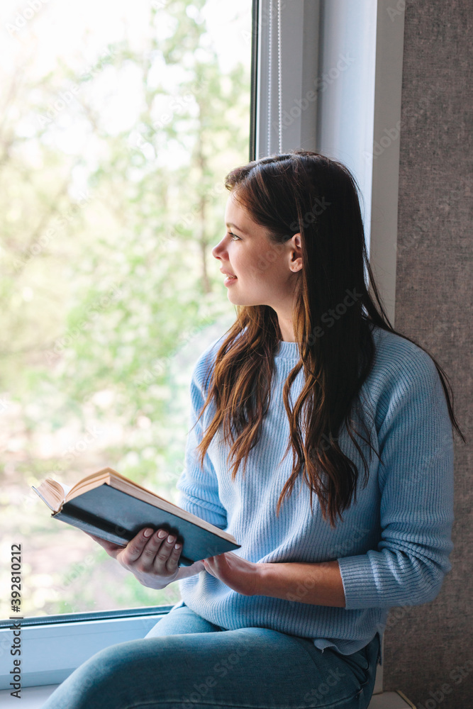 Fototapeta premium Young attractive woman reading book and looking at window