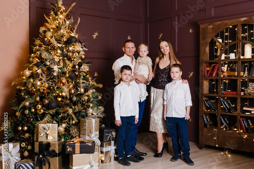 A beautiful happy young family of five stands in full height near the Christmas tree and look into the camera. mother, father and three children © Alyona