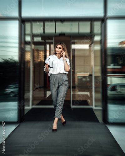 Vertical portrait of young woman small business owner comes out of an automatically opening door. Lady dressed in white bright shirt and pants hold mobile cell phone call speaking with client. 