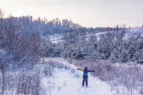 A girl in a ski suit on skis with her hands up in the fresh snow on the background of a snow-covered forest and the sky at sunset