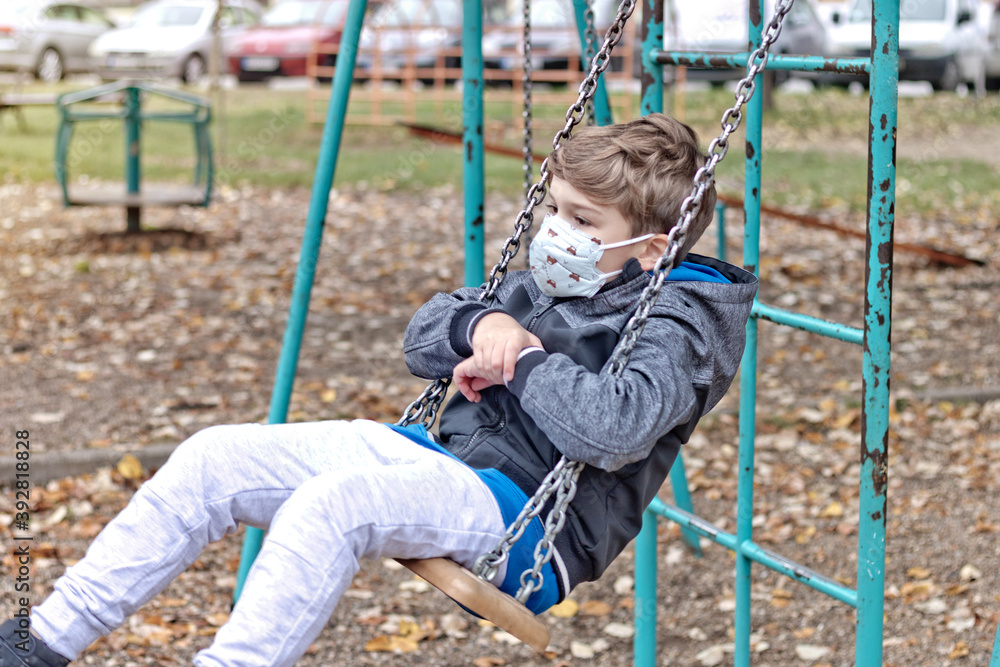 Pensive boy swinging on the playground while wearing face mask due to coronavirus pandemic. Pensive boy swinging on the playground while wearing face mask due to coronavirus pandemic.