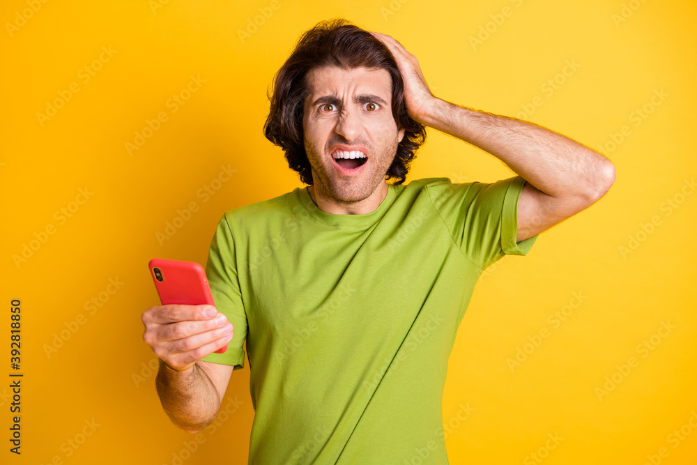 Photo of confused displeased guy hold cellphone arm head open mouth wear green t-shirt isolated yellow color background Photo of confused displeased guy hold cellphone arm head open mouth wear green t-shirt isolated yellow color background