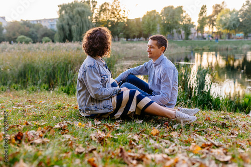 Fototapeta Naklejka Na Ścianę i Meble -  Loving couple hugging sitting on the bank of the river. A man hugs a round abdomen of his pregnant wife. The concept of paternity. Nine months. Expectation of the child. Place for text.