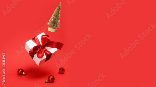 Holiday Christmas background. Golden winter tree, white gift box with red ribbon falling with balls and sparkling lights on reddish background for greeting card. Christmas, winter, new year concept.