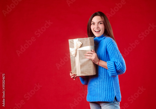 girl in a blue sweater holding a gift in her hands on a red background. © Ilnur
