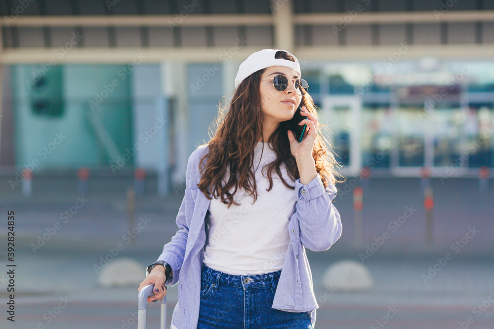 Young beautiful girl talking on the phone near a big bus stop with a suitcase