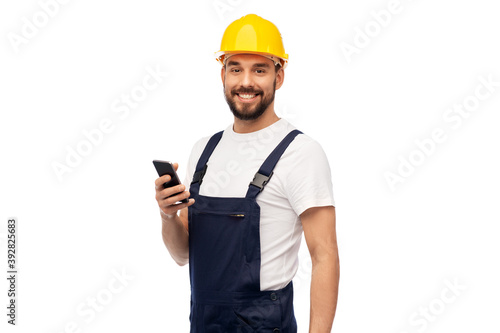 profession, construction and building - happy smiling male worker or builder in yellow helmet and overall with smartphone over white background