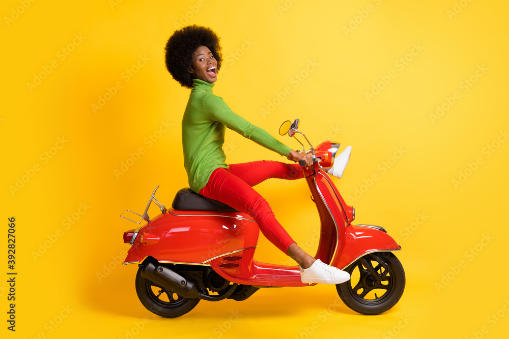 Fototapeta premium Photo portrait of excited african american woman driving a red scooter with legs spread wearing casual outfit isolated on vivid yellow colored background