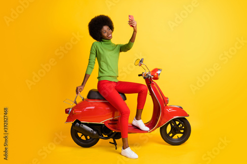 Photo portrait of young rider biker with brunette sitting on red retro motorbike holding cellphone taking selfie isolated on vivid yellow colored background