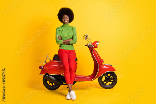 Photo portrait of young girl driver on red scooter with crossed hands isolated on vivid yellow colored background