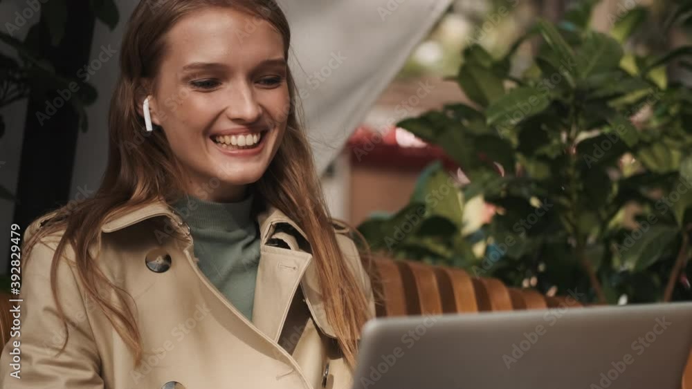 Attractive cheerful student girl in wireless earphones waving hello to friends during online lecture on laptop in cafe outdoor. Greeting expression Attractive cheerful student girl in wireless earphones waving hello to friends during online lecture on laptop in cafe outdoor. Greeting expression
