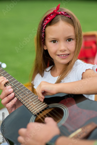 Portrait of happy little girl smiling at camera while sitting with her dad on green grass in park and playing with guitar